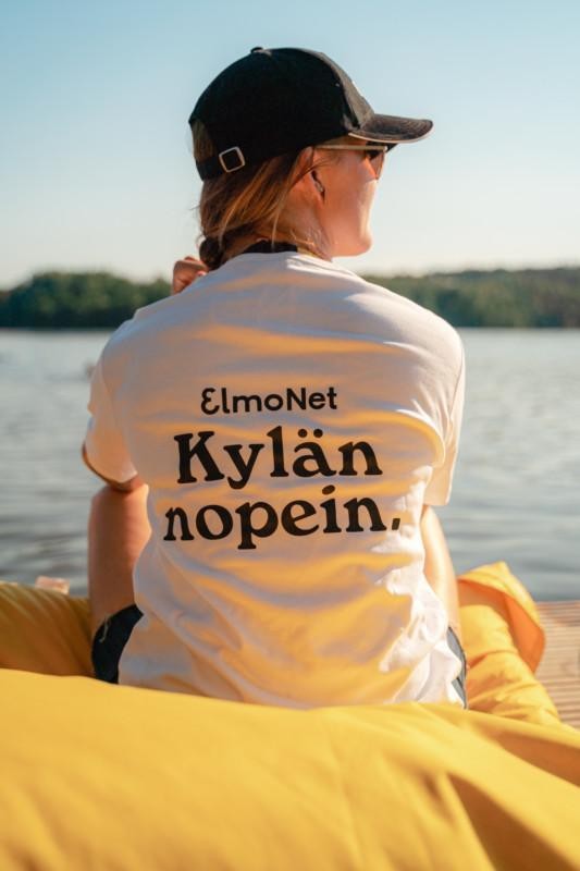 Nainen kesäauringossa säkkituolilla ElmoNetin Kylän nopein -paidassa. Kylän nopein netti myynnissä nyt myös Mänttä-Vilppulan Rusinniemessä.