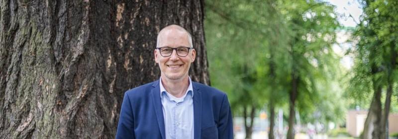 Silmälasipäinen mies puvuntakissa katsoo kameraan, taustalla puisto.