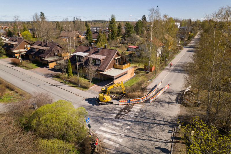 Keltainen kaivinkone työskentelee tienristeyksessä omakotitaloalueella.